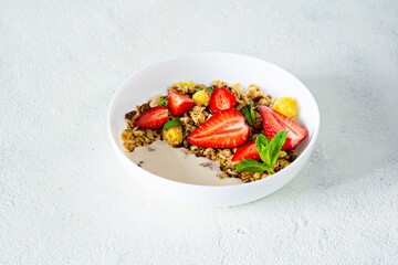 Homemade crunchy granola with nuts, dried fruits, fresh strawberries, mint and yogurt (fermented baked milk) on a white plate. Healthy breakfast concept.