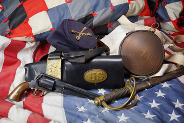 Fototapeta premium Artifacts from the American Civil War. Union army hat gear sit on top of an American flag and a patriotic quilt. A pistol and saber from the US Cavalry, canteen and leather items make up the display.