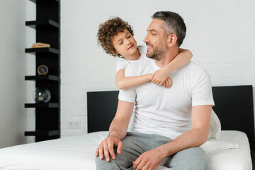 curly son hugging cheerful bearded father in bedroom