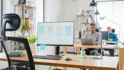 Desktop Computer Standing on the Desk in the Modern Creative Office. Screen Showing Smartphone Application UI / UX.  In the Background Bright Studio with Team of Young Professionals Work on Computers.