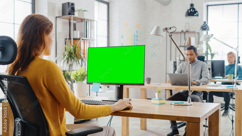 Poster over the shoulder: creative young woman sitting at her desk using desktop computer with mock-up gree