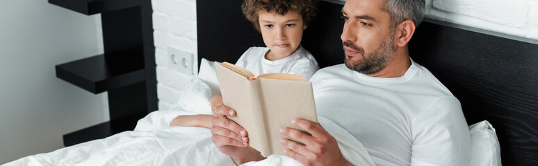 panoramic orientation of bearded father reading book near curly son in bedroom