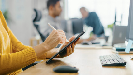 Fototapeta premium Anonymous Young Woman Sitting at Her Desk She's Drawing, Writing and Using Pen with Digital Tablet Computer. Focus on Hands with Pen. Bright Office where Diverse Team of Young Professionals Work.