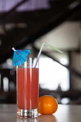 a tall glass with red-orange fresh juice, a straw and a blue cocktail umbrella on a light gray table in the interior. tangerine next to a glass