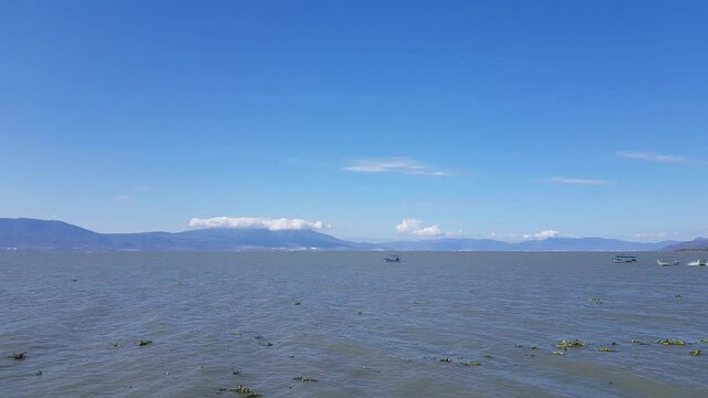 Slow shot of Chapala lake with gentle wind causing calm swell with seagulls flying, with mountains, the promenade and the city in the background, sunny day in Jalisco, Mexico