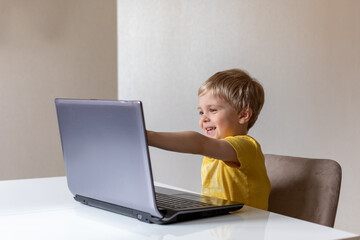 A cute little boy with blond hair sits at a table with a laptop and waves his hand. The boy smiles. He's got virtual communication.