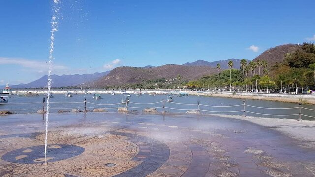 Fountain with a water jet on the boardwalk of Lake Chapala with anchored boats, the promenade with palm trees, it's gateway pier and gazebo, mountains in the background, sunny day in Jalisco, Mexico