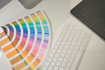 Woman freelance graphic designer at work in her home office chooses colors from palette for design and printing a new job. She work on a white desk with the black graphic tablet and computer keyboard.