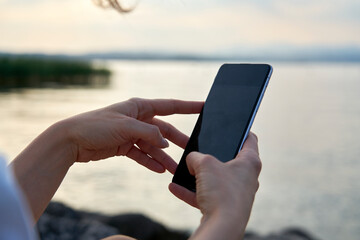 Pretty woman uses smartphone with her hands to photograph lake garda at sunset. The sun is reflected on the lake water and the sky is clear.