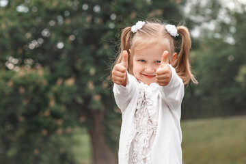 thumbs up. Blonde blue-eyed little girl in white on a background of green trees looks at the camera and shows thumbs up. Girl with two tails and white flowers on her head