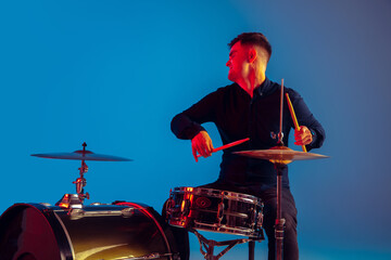 Caucasian male drummer improvising isolated on blue studio background in neon light. Performing,...