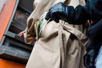 Low angle view of thief in leather gloves holding dollars near woman in coat using atm