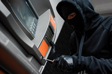 Low angle view of thief in balaclava using screwdriver while breaking atm
