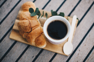 Cup of coffee on wooden background with croissant and white vase with greens. Breakfast or coffee brake theme.