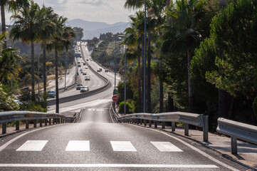 Obraz premium A minor road with palm trees along the roadsides adjoins a highway with cars, mountains in the background.