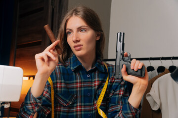 Portrait of a dressmaker girl looks at the sketch with contempt. A young seamstress holds a gun and a cigar. Craftswoman in a plaid shirt