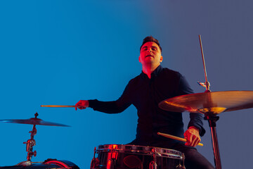 Caucasian male drummer improvising isolated on blue studio background in neon light. Performing,...