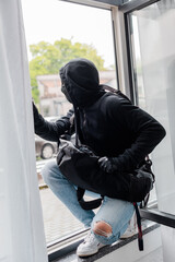 Selective focus of robber in balaclava and leather gloves holding bag while standing on window