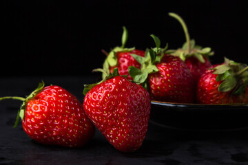 Strawberries on a black background. Two strawberries in the foreground. behind a plate with strawberries. Creative photo of strawberries