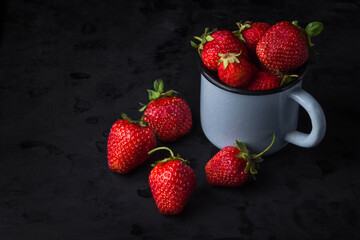 Strawberries on a black background. Enamel mug full of strawberries. Several strawberries lie nearby. Red strawberries. Creative photo of strawberries