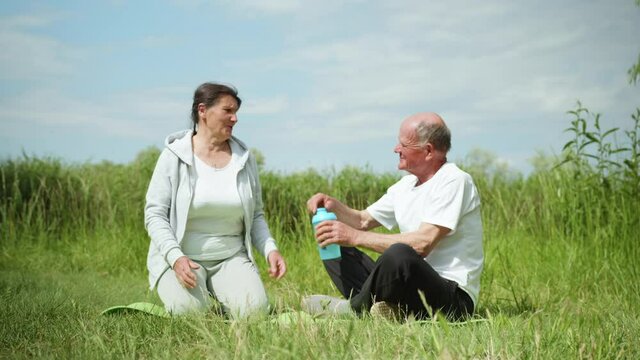 caring elderly man gives cool clean water in sports nutrition bottles to his dear beloved wife after playing sports or meditating, outdoors on background of green grass