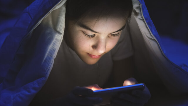 Toned portrait of smiling teenage girl lying in bed with smartphone and browsing internet - Powered by Adobe
