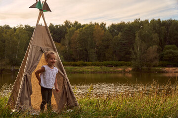A little girl is sitting by the lake. Sunny akat. Wigwam. Happy child. High quality photo