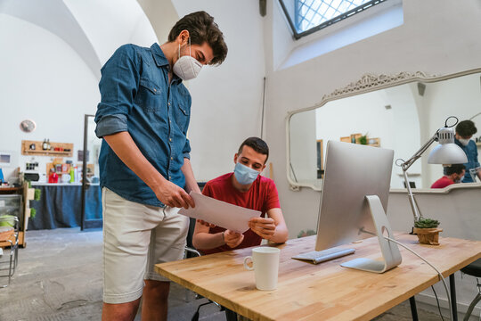 Two colleague millennials men at working with medical mask avoid the Coronavirus, Covid-19 discuss work situation analyzing graphs on performance of startup company for crisis due to the pandemic