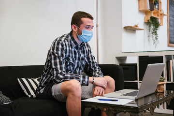 Young millennial man with shirt and shorts and medical mask in a video call conference at laptop on the sofa with his bosses of work at distance for avoid contamination from Coronavirus, Covid-19