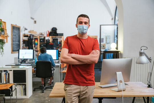 Portrait of leader of a startup in the office with medical mask for protection and prevention contagions from Coronavirus, Covid-19, behind him the other workstations with colleagues at computer
