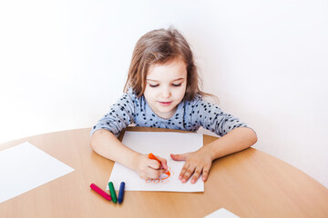 A little girl in a gray dress sits at a table and draws a flower with wax crayons, orange flower,...