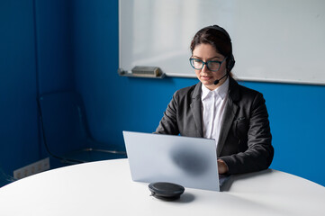 Serious woman wearing a headset is having an online conversation on a laptop in a conference room. Female boss communicates subordinates by video call. An employee is watching a business webinar.