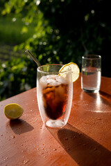 Iced beverage on glass with ice and lemon standing on the table in front of green field and tree