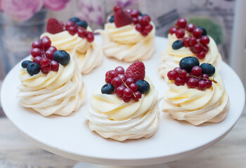 Six mini Pavlova meringue cakes with whipped cream, fresh red currant, raspberry and blueberry on white cakestand.