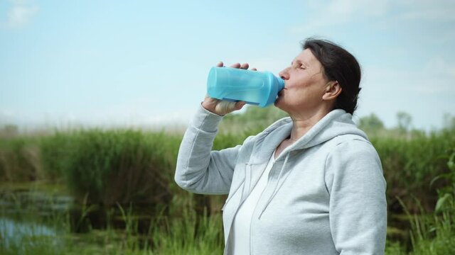 pretty old woman quenches thirst drinks water after doing sports outdoors in meadow by river on sunny summer day