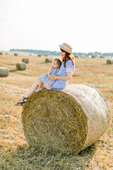Naklejka na ściany i meble Young beautiful woman in striped dress and straw hat sits on hay bale and hugs her adorable little baby girl, enjoying summer sunset in field with haystacks