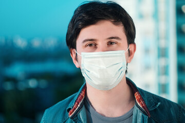 A young man in a medical mask blue and shirt stands looking ahead against the blurry backdrop of an city