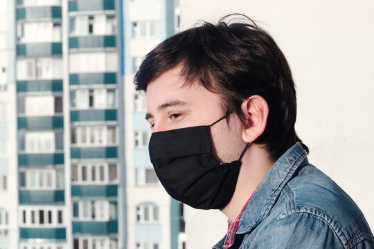 A young man in a medical mask black
looks into the distance on a balcony against the background of city buildings - Powered by Adobe