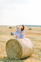 Naklejka na ściany i meble Happy two year old girl child in striped dress playing with young smiling mom in summer autumn field with hay bales, sitting together on hay stack. Summer sunset portrait