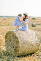 Naklejka na ściany i meble Happy cute little girl child in blue striped dress hugs her young mom, sitting toegethr on hay round bale in summer autumn wheat field