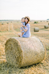 Naklejka na ściany i meble Happy cute little girl child in blue striped dress hugs her young mom, sitting toegethr on hay round bale in summer autumn wheat field