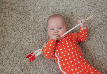 cupid and arrow, сute baby in a red bodysuit with white hearts lies on the carpet and holds an...