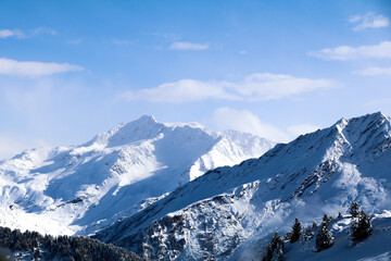 Beautiful jagged rocky mountains of the Alps with large depth of field. Trees lining the slope can be seen in the foreground covered in snow. Sunny blue-sky day.
