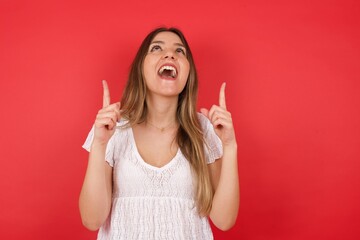 Charming carefree woman with positive expression, points up with both index fingers, dressed in casual clothing, has broad interested smile, isolated over gray background. Look there, please.