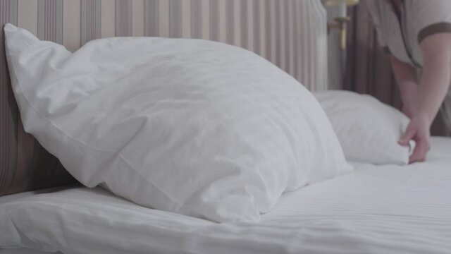 Close-up of pillow lying on big luxurious bed in hotel room with blurred employee putting cushion on bedding. Unrecognizable Caucasian maid working in tourist resort. Job, occupation, lifestyle.