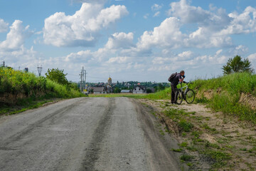 Dirt road to the village. A man with a backpack and a bicycle is standing on the roadside. On the horizon is a Ukrainian village with a church dome. Copy space.