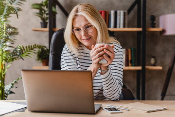 Portrait of happy senior woman using laptop while drinking coffee at workplace