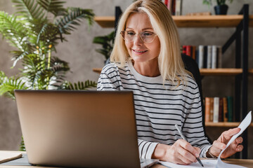 Senior stylish woman taking notes in notebook while using laptop at home office