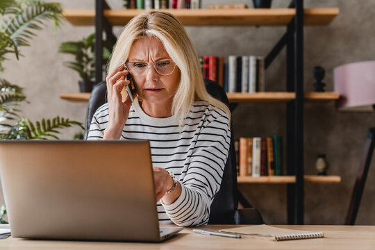 Mature woman looking stressed while using laptop and talking on mobile phone