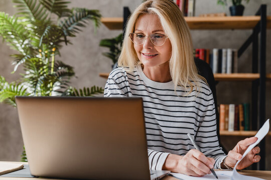 Senior Stylish Woman Taking Notes In Notebook While Using Laptop At Home Office